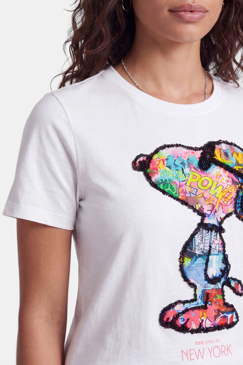 Eine Frau trägt ein weißes Snoopy-T-Shirt aus weicher Baumwolle mit einem farbenfrohen Snoopy-Motiv, das durch Pop-Art-inspirierte Comic-Elemente besticht und mit schwarzen Pailletten umrandet ist. Am unteren Rand des Shirts steht in rosa Schrift „see you in NEW YORK“. Zu sehen sind nur ihr Oberkörper und ein Teil ihres Gesichts.