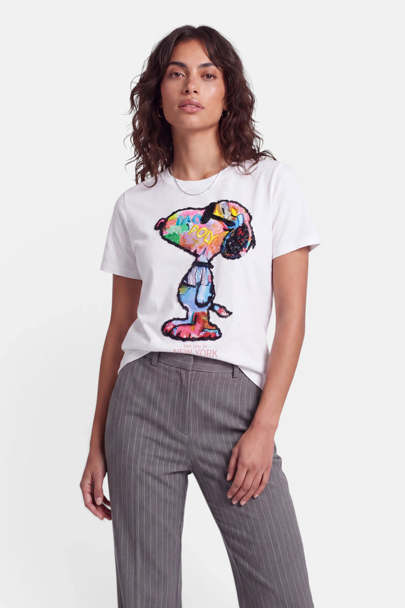 Eine Frau mit welligem braunem Haar trägt ein weißes Baumwolle-Snoopy-T-Shirt mit einer farbenfrohen, von der Pop-Art inspirierten abstrakten Snoopy-Grafik sowie eine graue Nadelstreifenhose. Sie steht vor einem schlichten weißen Hintergrund, blickt mit neutralem Gesichtsausdruck in die Kamera und hat eine Hand in der Tasche.