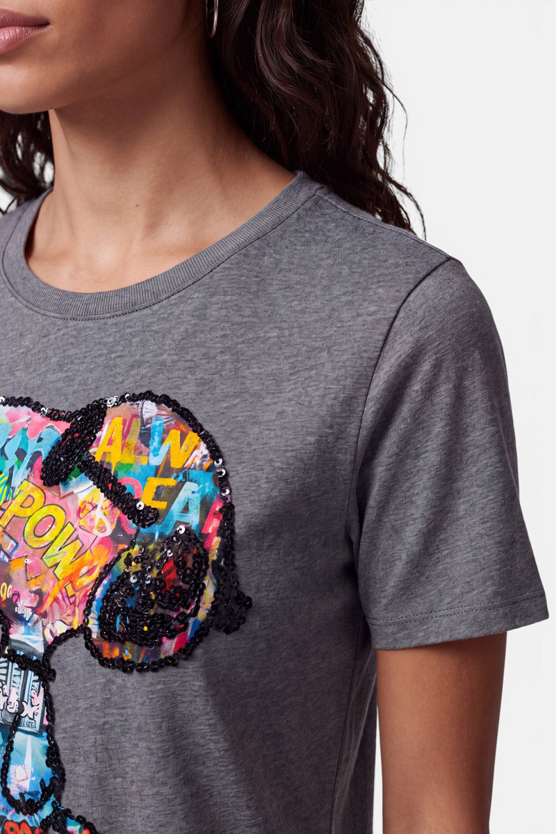 Eine Frau mit welligem, dunklem Haar trägt ein Snoopy-T-Shirt aus 100 % Baumwolle, das mit einer farbenfrohen, mit Pailletten verzierten Snoopy-Grafik mit Comic-Text und lebhaften Pop-Art-Mustern verziert ist, die sich vom Hals bis zum Oberarm vor einem schlichten weißen Hintergrund abheben.