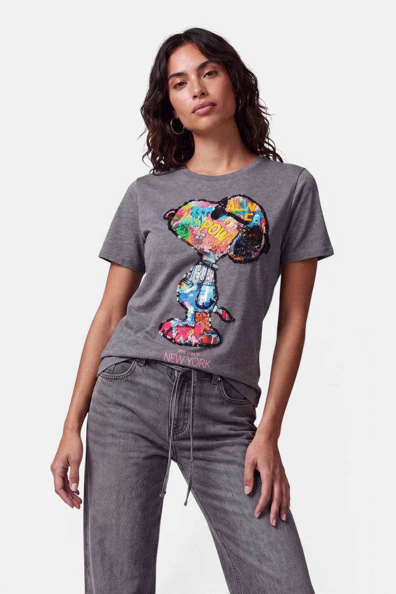 Eine Frau mit welligem, dunklem Haar trägt ein graues Snoopy-T-Shirt aus 100 % Baumwolle mit einem farbenfrohen Pop-Art-Print und dem Schriftzug „NEW YORK“ in kräftiger Schrift am Saum. Dazu trägt sie eine graue Jeans und steht vor einem schlichten weißen Hintergrund, während sie selbstbewusst in die Kamera blickt.