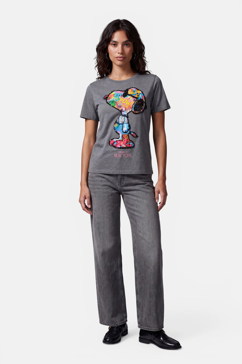 Eine Frau mit welligem, dunklem Haar trägt ein graues Snoopy-T-Shirt aus 100 % Baumwolle mit einem farbenfrohen Pop-Art-Print im Graffiti-Stil und dem Schriftzug „NEW YORK“. Dazu trägt sie eine graue Jeans mit weitem Bein und schwarze Schuhe und steht vor einem schlichten weißen Hintergrund.