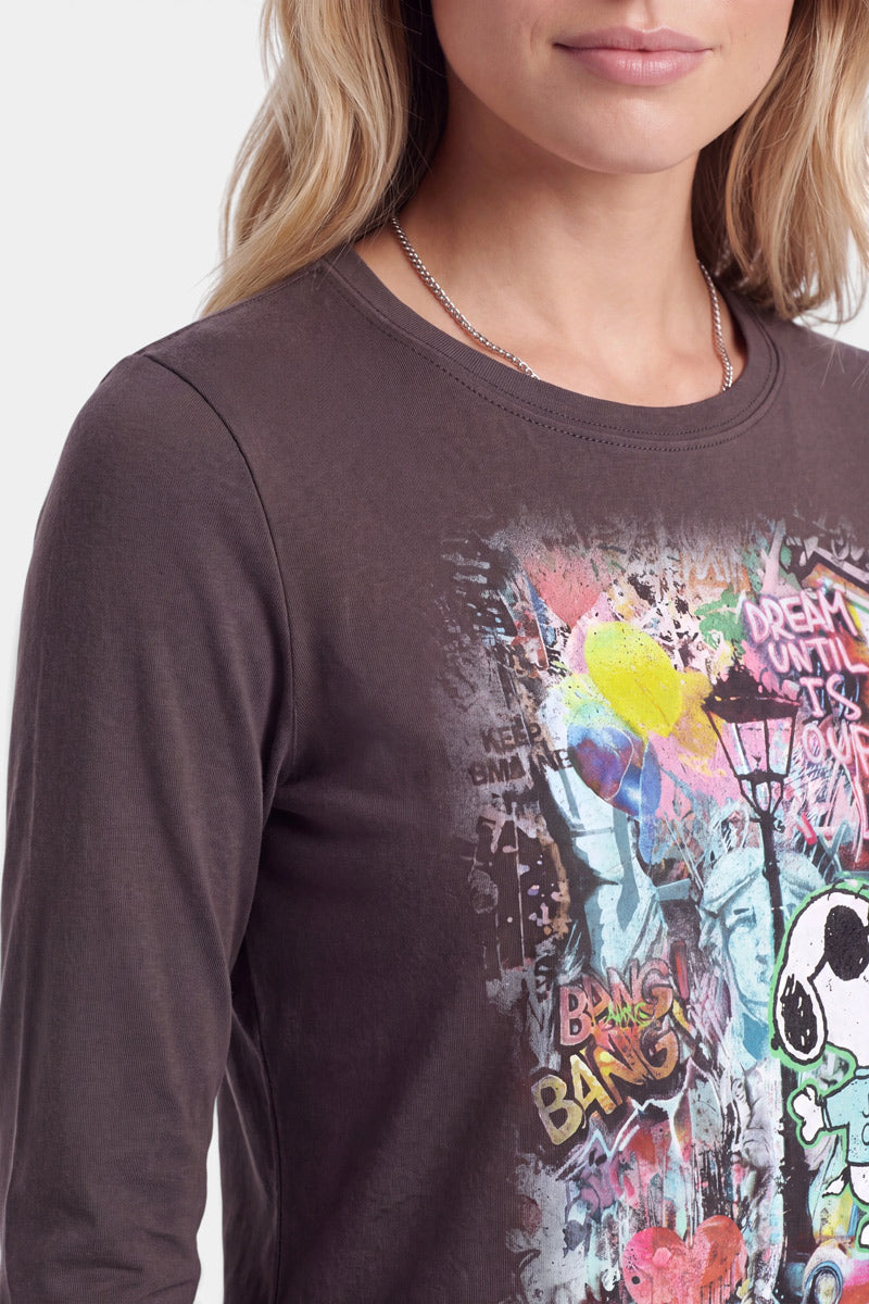 Eine Frau mit langen blonden Haaren und hellem Lippenstift trägt ein dunkelgraues Langarmshirt mit farbenfrohen Motiven im Graffiti-Stil, die von „Peanuts“-Kunst inspiriert sind, darunter Comicfiguren wie Snoopy im Pop-Art-Stil, Farbspritzer, eine Straßenlaterne und Schriftzüge. Nur ihr Kopf und ihr Oberkörper sind zu sehen.