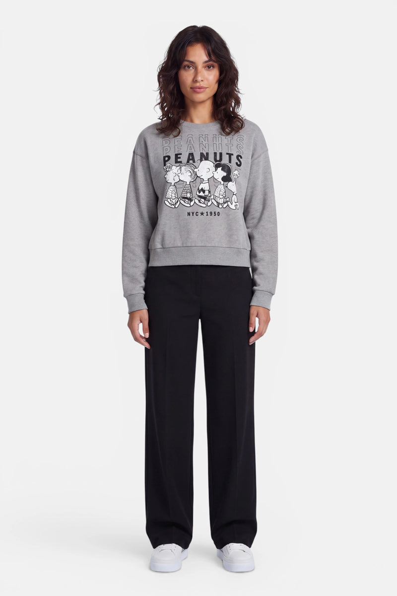 Eine Frau steht mit dem Blick nach vorne und trägt ein graues „Peanuts“-Sweatshirt aus Baumwolle mit einem verspielten Aufdruck, eine lockere schwarze Hose und weiße Sneaker. Sie hat welliges dunkles Haar, einen neutralen Gesichtsausdruck und steht vor einem schlichten weißen Hintergrund.