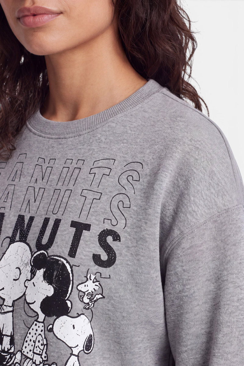 Eine Frau mit welligem braunem Haar trägt ein hellgraues „Peanuts“-Sweatshirt, auf dem Comicfiguren und der Schriftzug „PEANUTS“ in fetter schwarzer Schrift zu sehen sind. Das Bild ist aus weicher Baumwolle gefertigt und so beschnitten, dass sie von unterhalb der Augen bis zur Brust zu sehen ist, wobei der Fokus auf dem Design des Sweatshirts liegt.