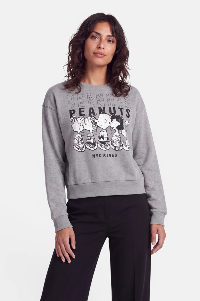 Eine Frau mit welligem braunem Haar trägt ein graues „Peanuts“-Sweatshirt aus Baumwolle mit Comicfiguren und eine schwarze Hose. Sie steht vor einem schlichten weißen Hintergrund, blickt mit entspanntem Gesichtsausdruck in die Kamera und hält eine Hand an der Seite.