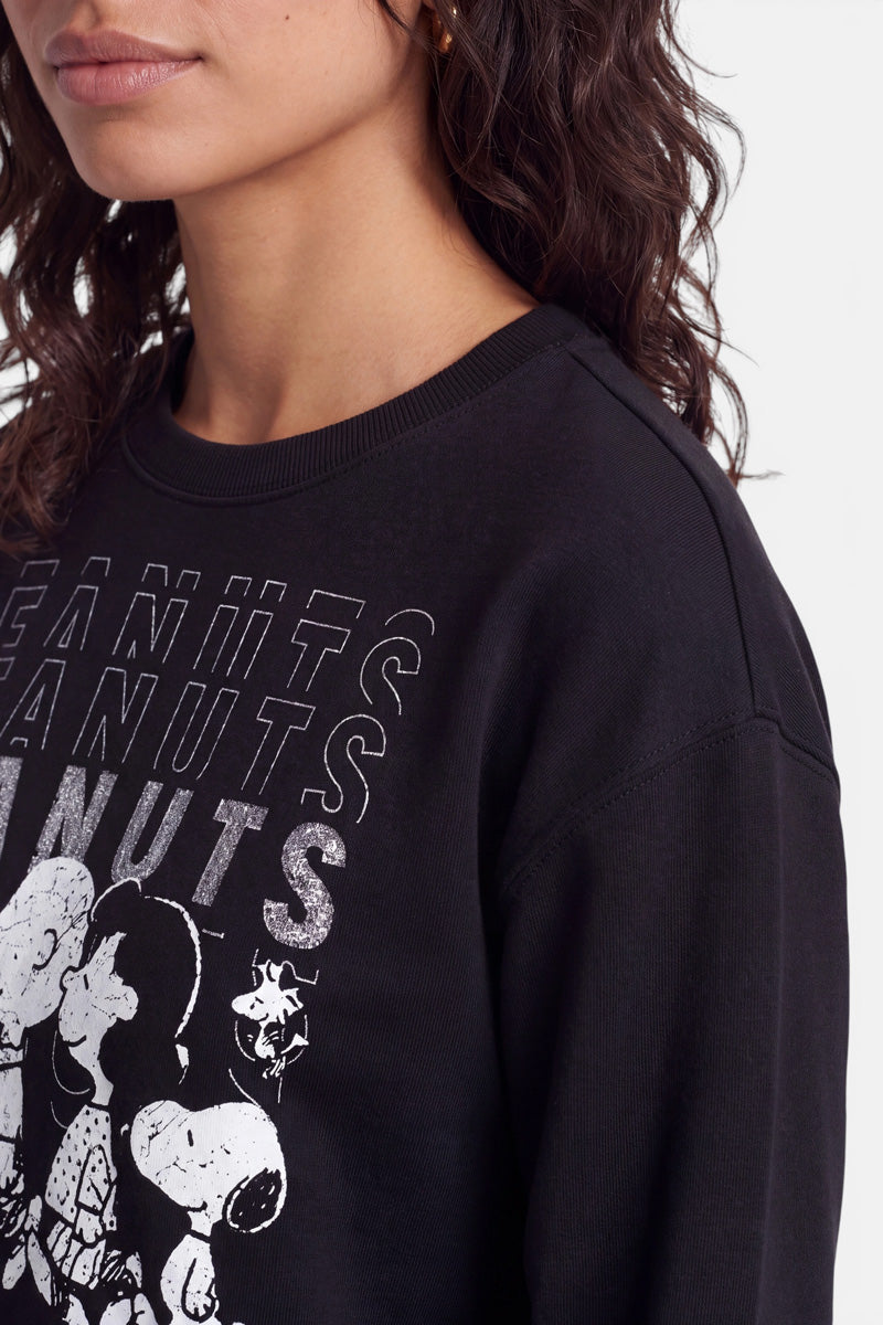 Eine Frau mit welligem, dunklem Haar trägt ein schwarzes „Peanuts“-Sweatshirt aus weicher Baumwolle. Das Sweatshirt ist mit verblassten Abbildungen von Snoopy und Charlie Brown sowie einem sich wiederholenden weißen „PEANUTS“-Schriftzug verziert. Sie blickt leicht nach links; nur der untere Teil ihres Gesichts und ihres Oberkörpers sind zu sehen.