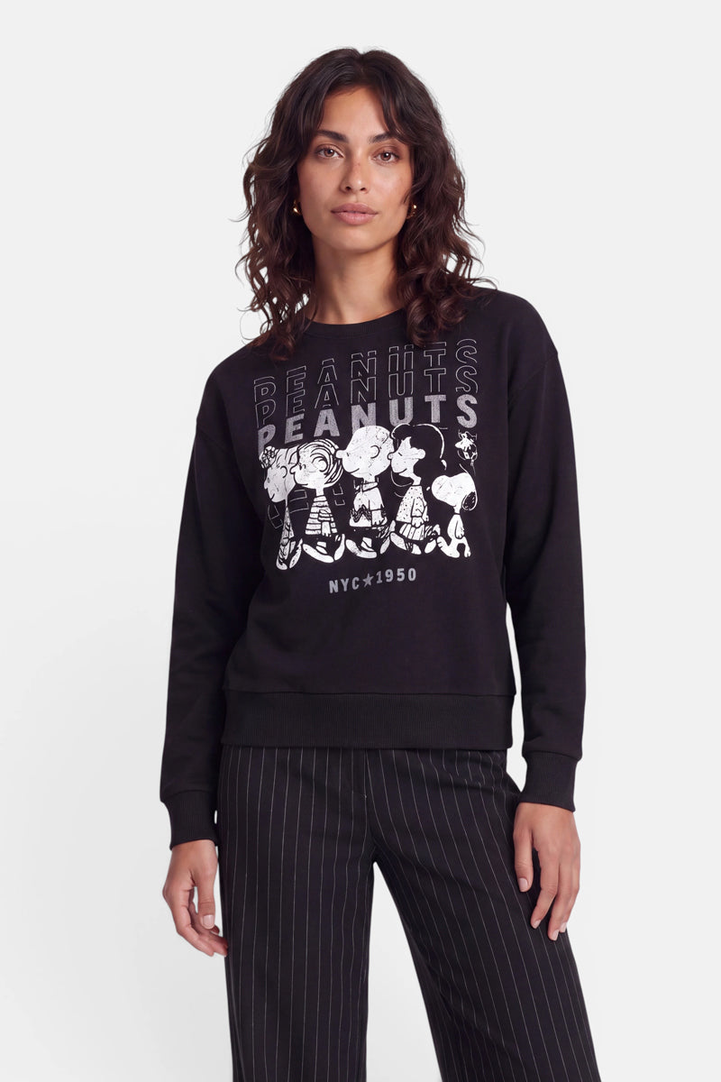 Eine Frau mit welligem braunem Haar trägt ein schwarzes „Peanuts“-Sweatshirt aus Baumwolle mit weißen Comicfiguren und Schriftzügen sowie eine schwarze Nadelstreifenhose. Sie steht mit dem Gesicht nach vorne vor einem schlichten weißen Hintergrund und hat einen neutralen Gesichtsausdruck.