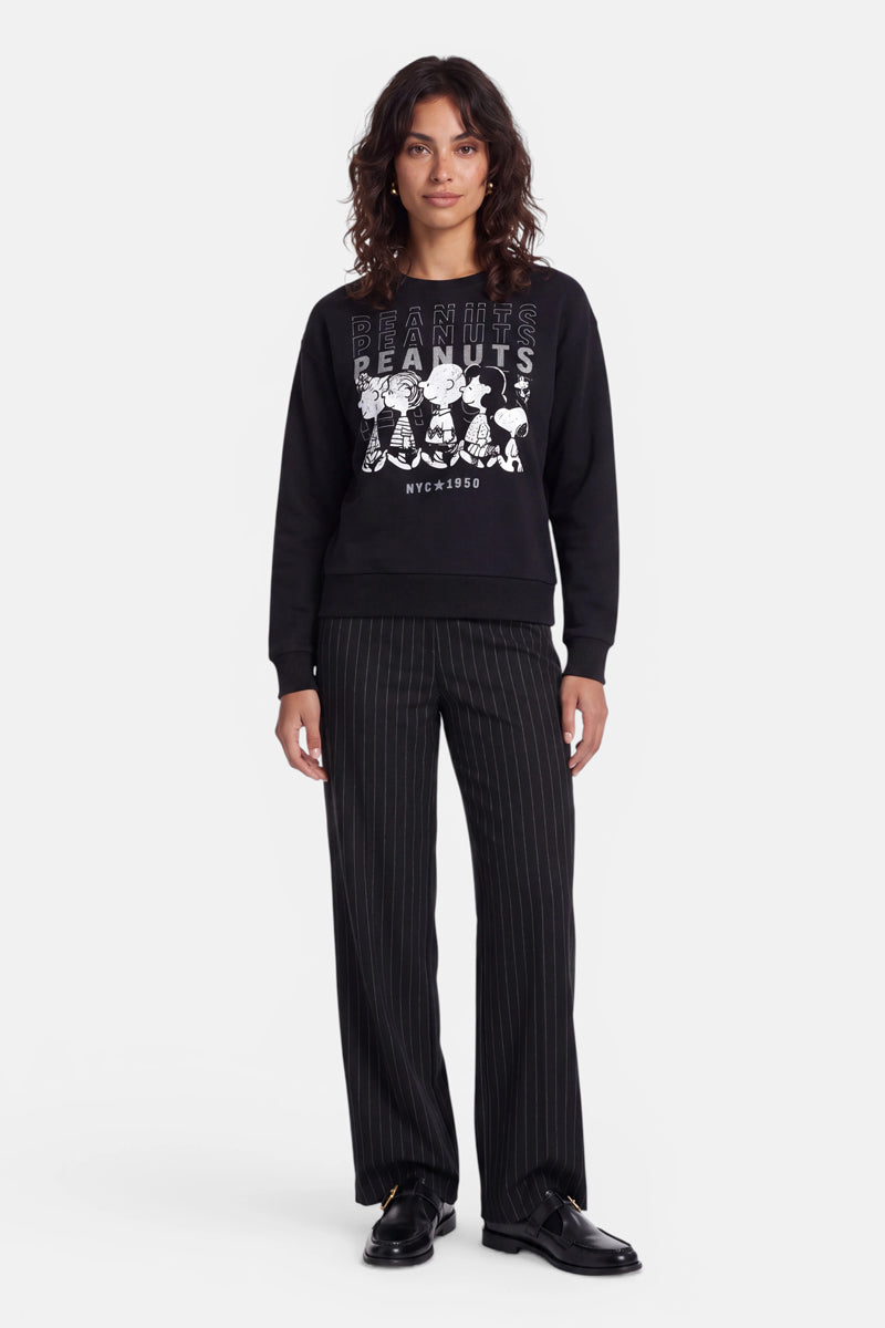 Eine Frau mit welligem, dunklem Haar trägt ein schwarzes „Peanuts“-Sweatshirt aus weicher Baumwolle mit Comicfiguren darauf, dazu eine schwarze Nadelstreifenhose und schwarze Schuhe. Sie steht mit dem Blick nach vorne vor einem schlichten weißen Hintergrund, in einer entspannten Haltung und mit einem leichten Lächeln.