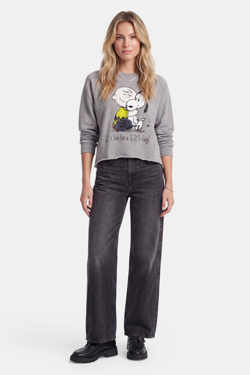 Eine Frau mit langen blonden Haaren trägt ein graues Snoopy-Sweatshirt aus Baumwolle, eine weite schwarze Jeans und schwarze Schuhe. Sie steht mit dem Blick nach vorne vor einem schlichten weißen Hintergrund, mit neutralem Gesichtsausdruck und den Armen entspannt an den Seiten.