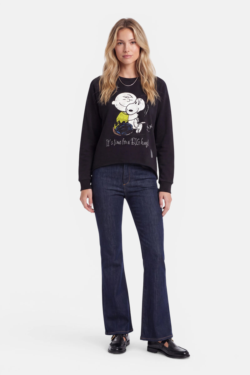 Eine Frau steht mit dem Blick nach vorne und trägt ein schwarzes Snoopy-Sweatshirt aus 100 % Baumwolle, dunkelblaue Schlagjeans und schwarze Schuhe. Ihr langes blondes Haar trägt sie offen. Das Sweatshirt mit Rundhalsausschnitt zeigt Snoopy, wie er Woodstock umarmt, sowie den Text „It’s time for a BIG hug!“. Der Hintergrund ist schlicht weiß.