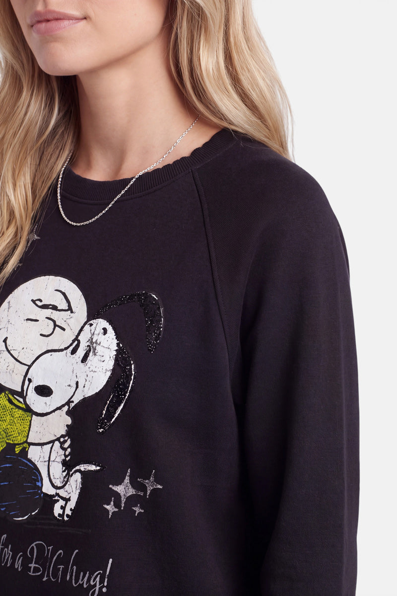 Eine Frau mit blonden Haaren trägt ein kuscheliges Snoopy-Sweatshirt aus weicher Baumwolle, auf dem Snoopy abgebildet ist, wie er Charlie Brown umarmt. Außerdem trägt sie eine dünne silberne Halskette. Der Hintergrund ist einfarbig und hellgrau. Nur die untere Hälfte ihres Gesichts und ihres Oberkörpers sind zu sehen.