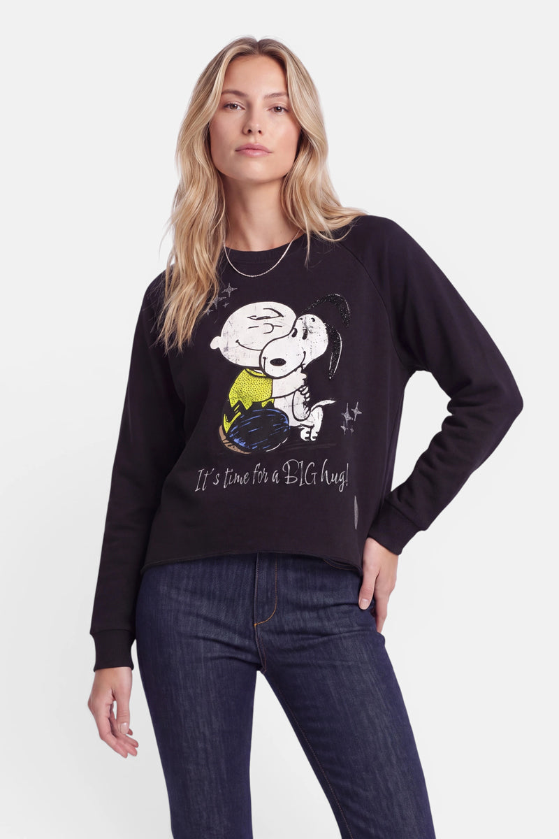 Eine Frau in dunklen Jeans und einem Snoopy-Sweatshirt aus weicher Baumwolle, auf dem ein „Peanuts“-Comic-Motiv zu sehen ist, bei dem Charlie Brown Snoopy umarmt. Der Text lautet: „Es ist Zeit für eine GROSSE Umarmung!“ Sie steht mit einer Hand in der Hüfte vor einem weißen Hintergrund.