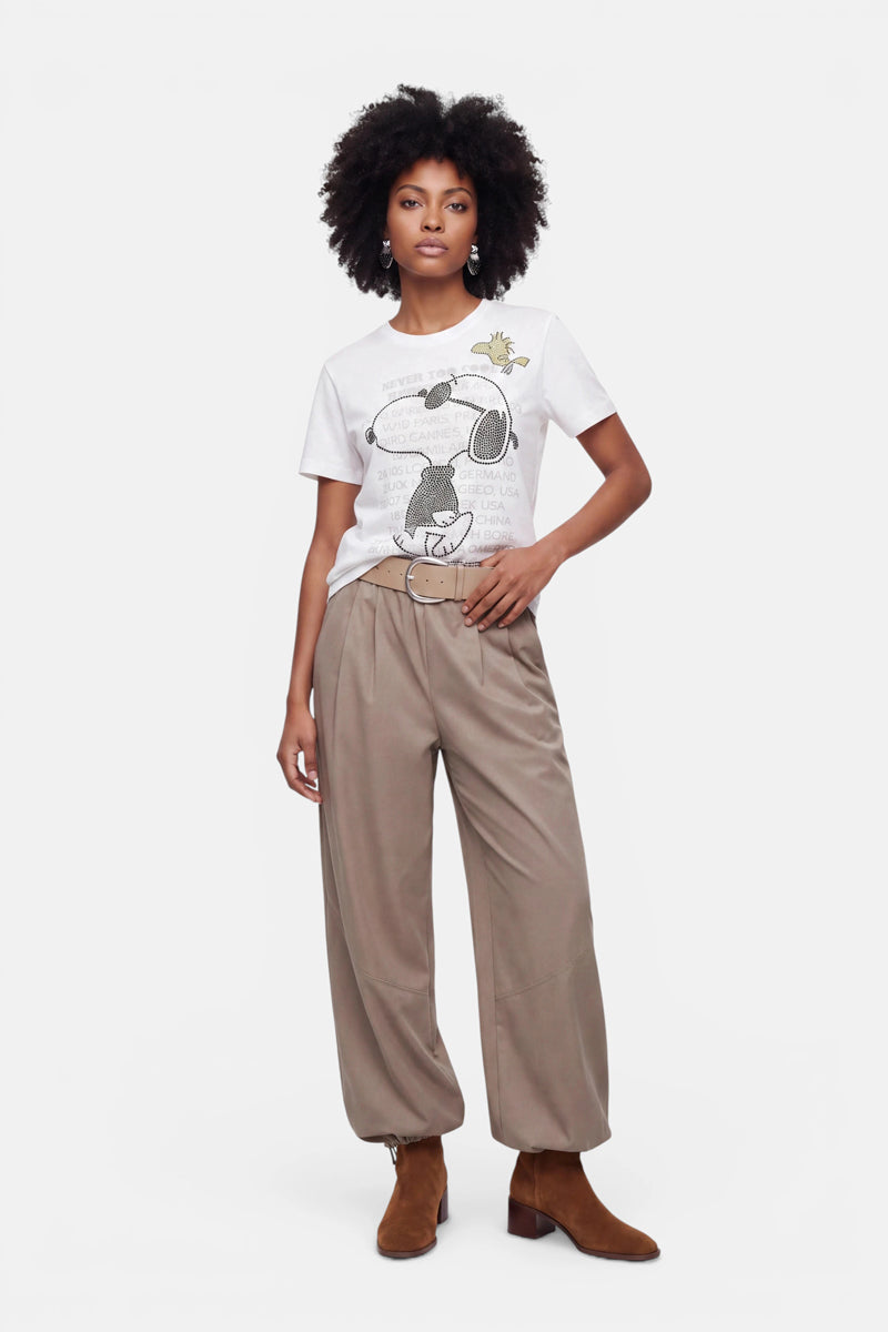 Eine Frau mit natürlich lockigem Haar trägt ein weißes Snoopy-T-Shirt aus Baumwolle, dazu eine lockere, hellbraune Hose mit hoher Taille und Gürtel sowie braune Wildleder-Stiefeletten. Sie steht vor einem schlichten weißen Hintergrund und hat eine Hand in der Tasche.