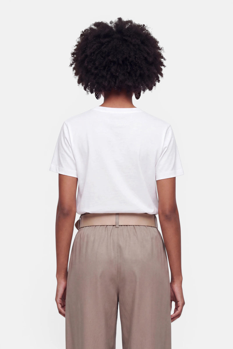 Eine Person mit kurzen, lockigen Haaren ist von hinten zu sehen. Sie trägt ein schlichtes weißes Baumwoll-T-Shirt, das in eine hellbraune Hose mit hoher Taille und Gürtel gesteckt ist. Der Hintergrund ist schlicht weiß.