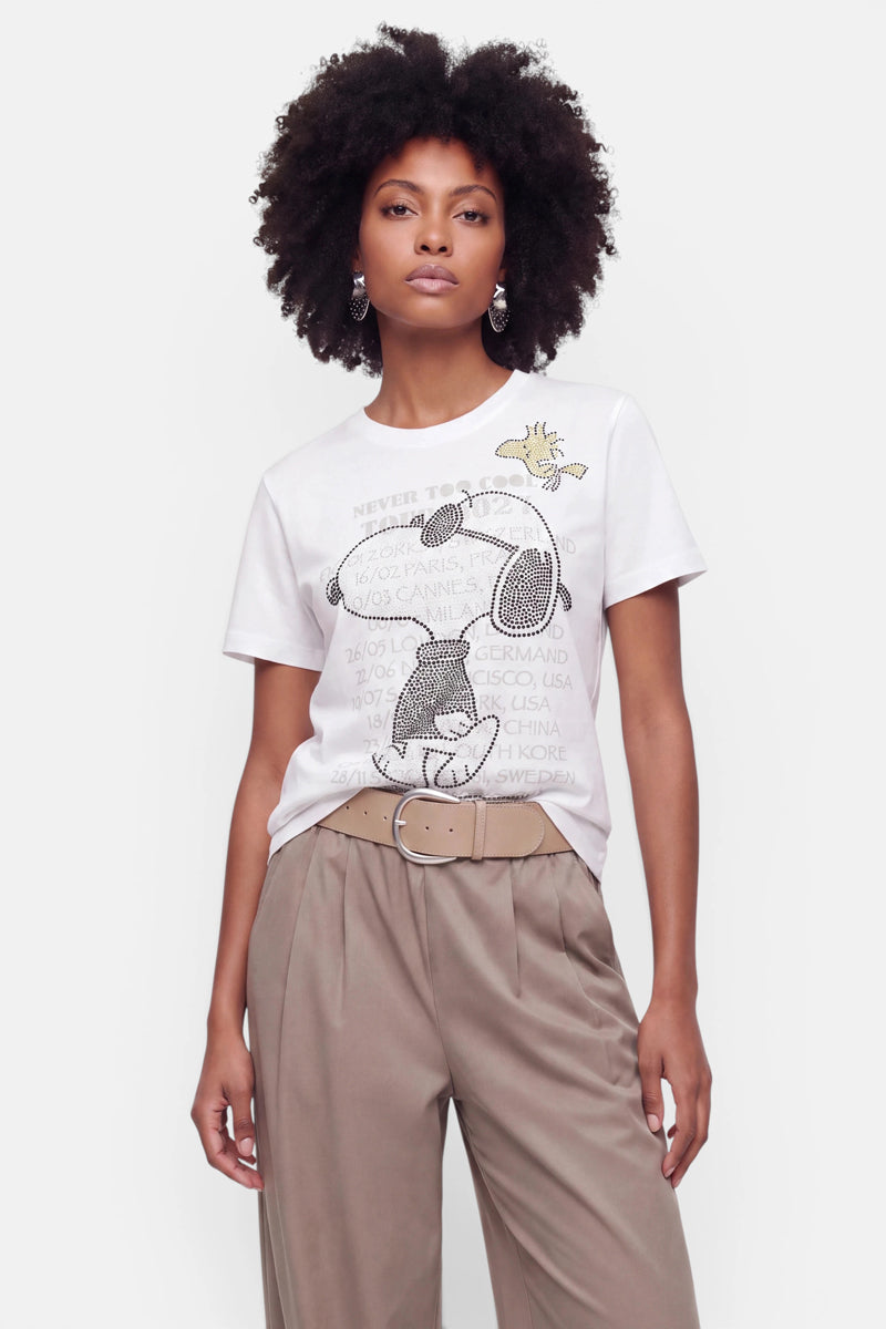 Eine Frau mit natürlich lockigem Haar trägt ein weißes Snoopy-T-Shirt aus Baumwolle mit dem beliebten Motiv und einem Schriftzug, das sie in eine beige Plisseehose mit hoher Taille und Gürtel gesteckt hat. Sie steht vor einem schlichten weißen Hintergrund, blickt in die Kamera und trägt dazu Hängeohrringe.