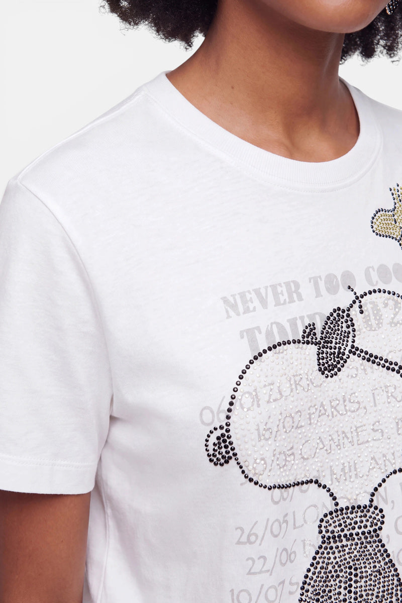 Eine Person trägt ein weißes Snoopy-T-Shirt aus Baumwolle mit einem schwarz-silbernen Strass-Motiv, das Snoopy und Woodstock zeigt. Auf dem Shirt sind teilweise lesbare Texte zum Thema Reisen zu sehen, darunter Städtenamen und Daten. Das Gesicht der Person ist teilweise außerhalb des Bildausschnitts.