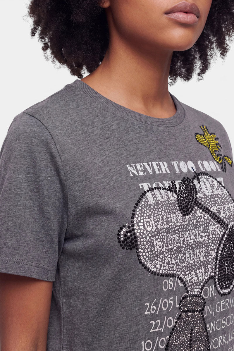 Eine Person, die ein graues Snoopy-T-Shirt aus Baumwolle trägt, das mit einer Strassverzierung versehen ist und Motive von Snoopy und Woodstock sowie weiße Tourdaten und den Schriftzug „NEVER TOO COOL TO ROCK“ zeigt. Die Person hat lockiges Haar, und auf dem Bild ist nur die untere Hälfte ihres Gesichts zu sehen.