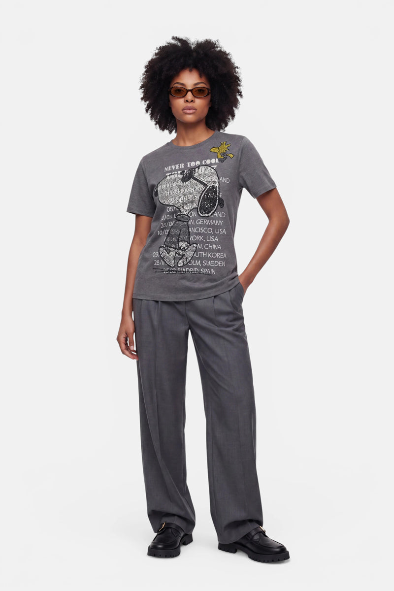 Eine Frau mit kurzen Locken und Sonnenbrille trägt ein graues Snoopy-T-Shirt aus Baumwolle mit Rundhalsausschnitt, auf dem ein Schriftzug und eine Margerite abgebildet sind, dazu eine graue Hose mit weitem Bein und schwarze Loafer. Sie steht mit dem Blick nach vorne und einer Hand in der Tasche vor einem schlichten weißen Hintergrund.