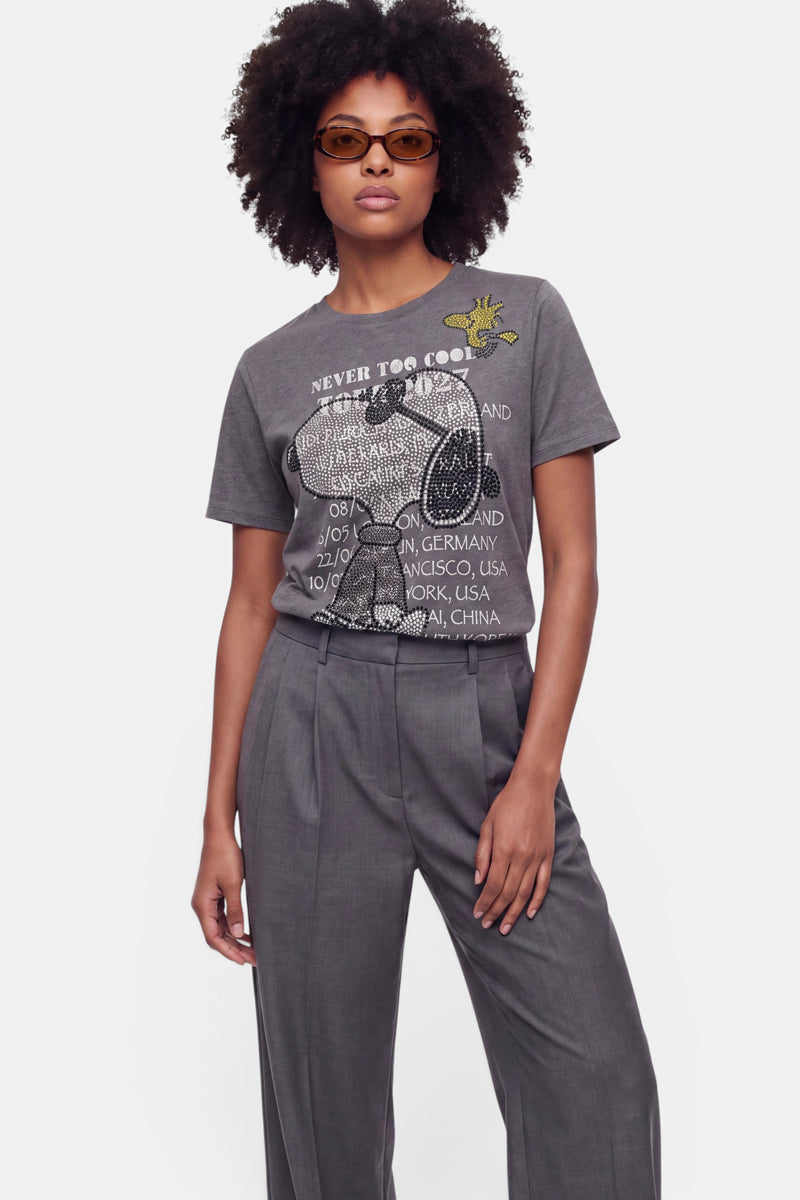 Eine Frau mit lockigem Haar trägt eine braune Sonnenbrille und ein graues Snoopy-T-Shirt mit Rundhalsausschnitt, auf dem ein Schriftzug und eine Comic-Hundefigur zu sehen sind. Dazu trägt sie eine graue Hose mit hoher Taille aus weicher Baumwolle und steht selbstbewusst vor einem schlichten weißen Hintergrund, die linke Hand in der Tasche.