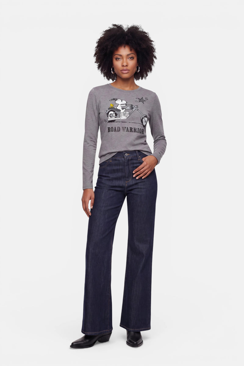 Eine Frau steht mit dem Blick nach vorne und trägt ein graues Langarmshirt aus Baumwolle mit einem „ROAD WARRIOR“- und Snoopy-Aufdruck, eine dunkle Jeans mit weitem Bein und schwarze Stiefel. Sie hat lockiges Haar und einen ausdruckslosen Gesichtsausdruck vor einem schlichten weißen Hintergrund.