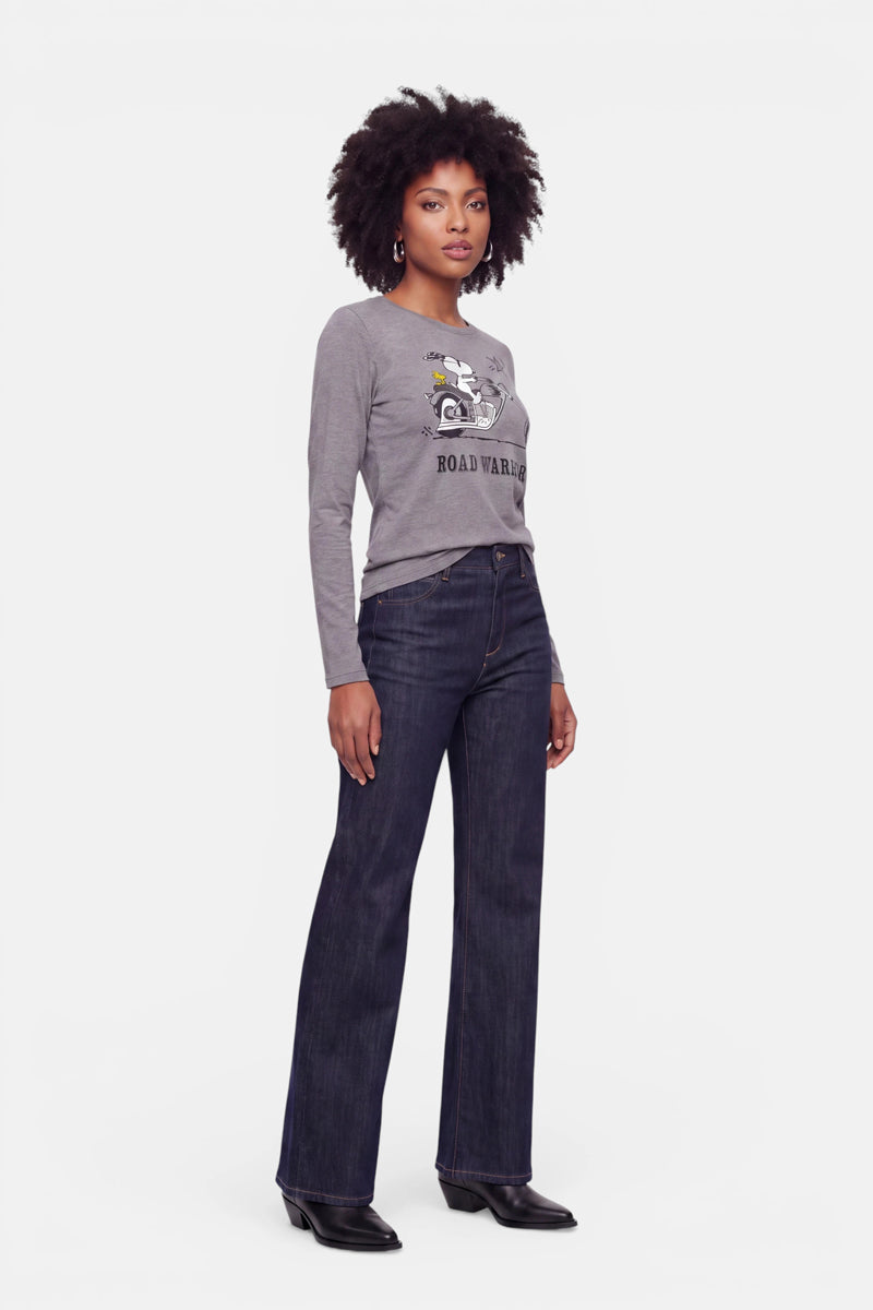 Vor einem weißen Hintergrund steht eine Frau mit dunklem, lockigem Haar. Sie trägt ein graues Langarmshirt aus Baumwolle mit Snoopy-Aufdruck, eine dunkle, hoch taillierte Schlagjeans und schwarze Stiefel. Sie blickt mit ausdruckslosem Gesicht geradeaus und hält die Arme an den Seiten.