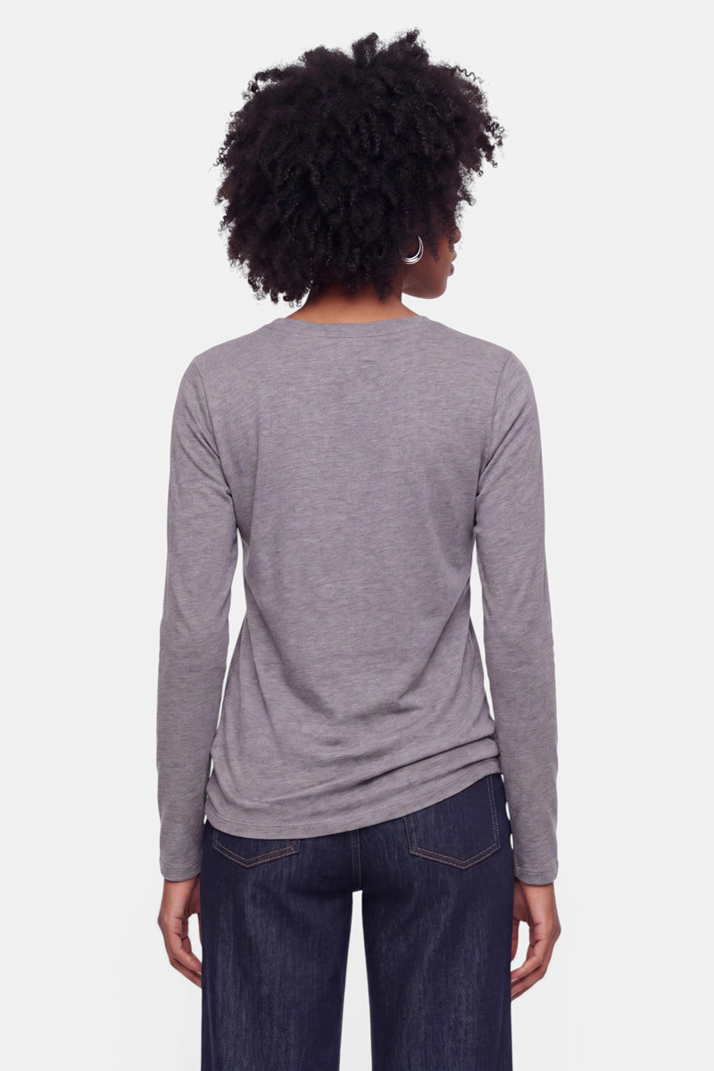 Eine Person mit kurzen Locken, die Creolen trägt, ein graues Baumwoll-Langarmshirt mit einem dezenten Snoopy-Aufdruck und dunkelblaue Jeans, steht mit dem Rücken zur Kamera vor einem schlichten weißen Hintergrund.