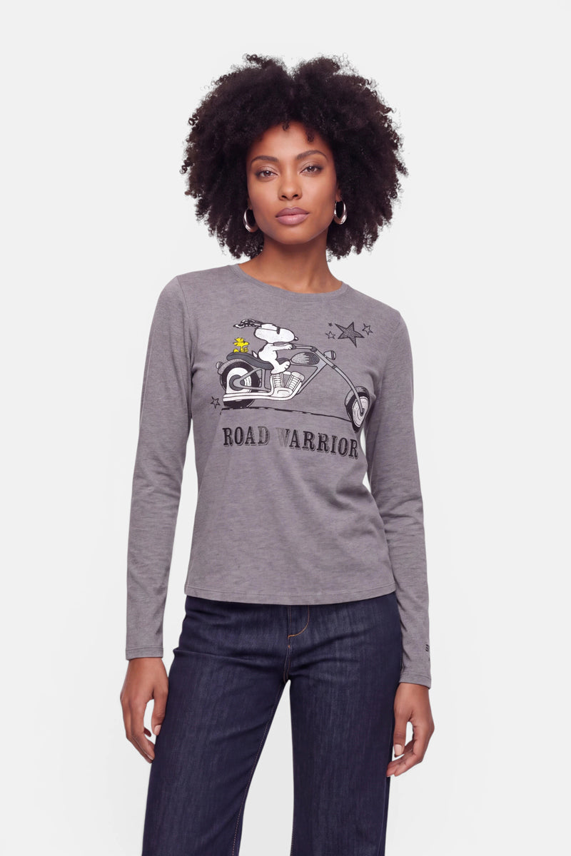 Eine Frau mit lockigem Haar trägt ein graues Langarmshirt aus Baumwolle mit einem Snoopy-Aufdruck, auf dem er auf einem Motorrad sitzt, sowie Sternen und der Aufschrift „ROAD WARRIOR“. Sie steht mit dem Blick nach vorne vor einem schlichten weißen Hintergrund und trägt eine dunkle Jeans sowie Creolen.