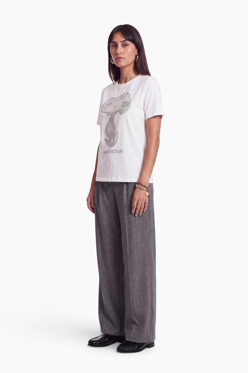 Eine Frau steht im Profil und trägt ein Snoopy-T-Shirt aus Baumwolle mit der Aufschrift „HAVE A NICE DAY“, dazu eine graue Hose und schwarze Schuhe.