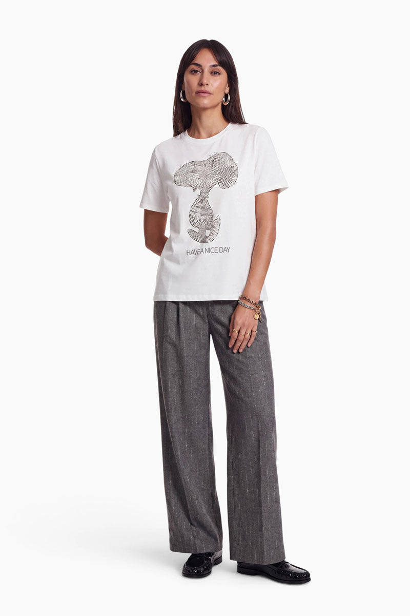 Eine Frau trägt ein weißes Baumwoll-T-Shirt mit einem mit Strasssteinen verzierten Snoopy-Motiv und dem Schriftzug „HAVE A NICE DAY“. Dazu trägt sie eine anthrazitgraue Hose mit weitem Bein und schwarze Loafer.