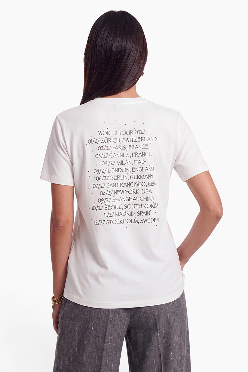Eine Person, von hinten gesehen, die ein weißes Snoopy-T-Shirt aus Baumwolle trägt. Auf der Rückseite des Shirts ist eine Liste der Orte und Termine der World Tour 2027 abgebildet, die von Zürich bis Stockholm reicht.