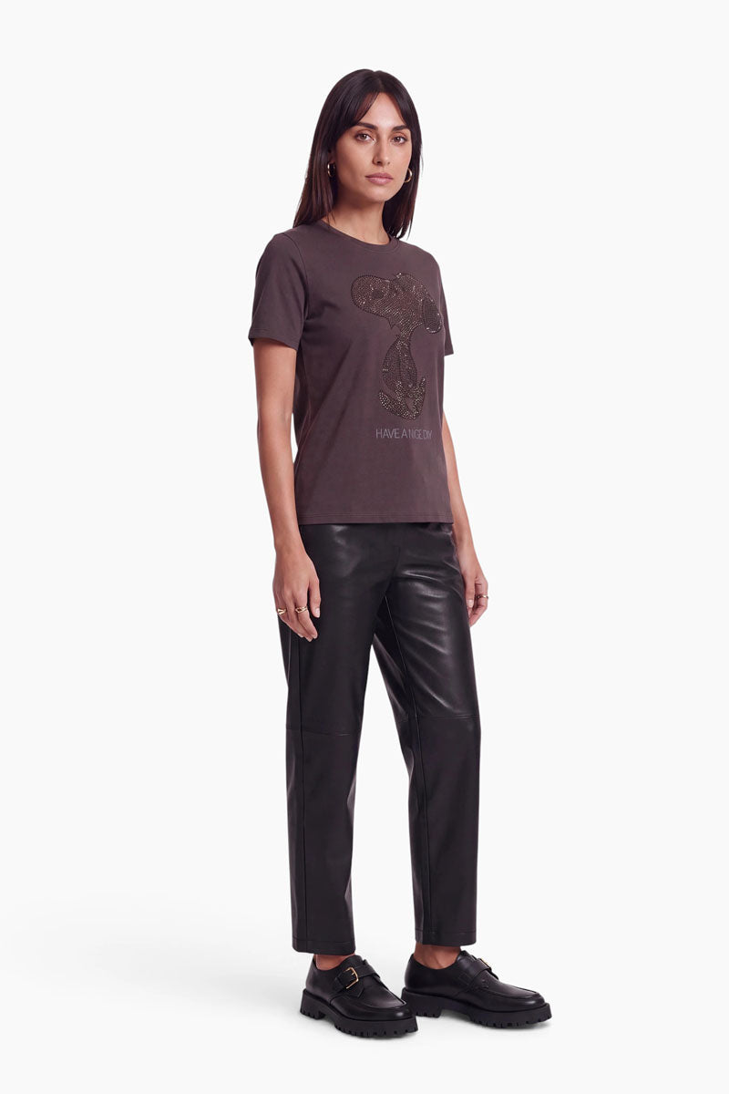 Eine Frau, die ein dunkelbraunes Baumwoll-T-Shirt mit einem aus Nieten geformten Snoopy-Motiv, eine schwarze Hose und schwarze Schuhe mit Schnallen trägt. Unter dem Snoopy-Motiv ist der Schriftzug „HAVE A NICE DAY“ aufgedruckt.