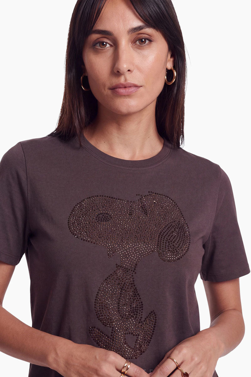 Eine Frau trägt ein braunes, kurzärmeliges Snoopy-T-Shirt aus Baumwolle, auf dem die Figur aus dunklen, reflektierenden Nieten abgebildet ist.