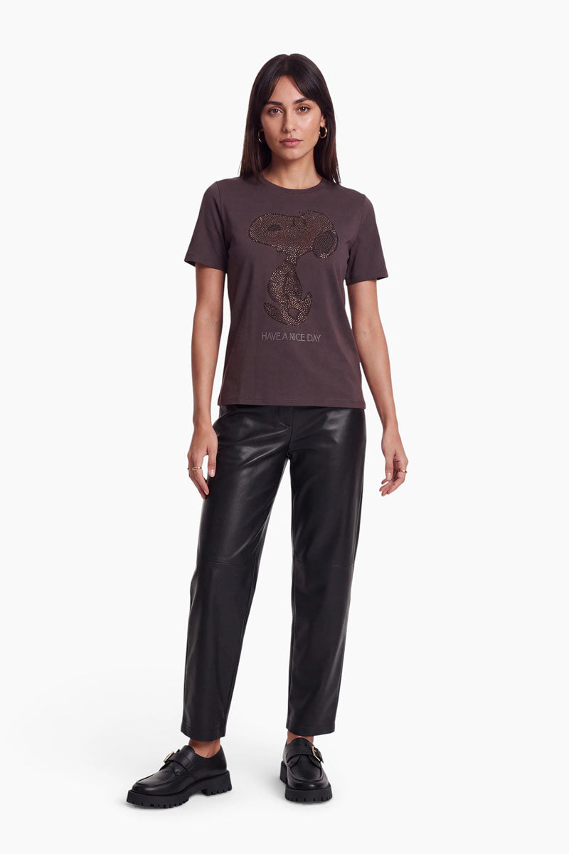 Eine Frau trägt ein braunes Snoopy-T-Shirt aus Baumwolle mit einer aus Perlen gestickten Grafik über dem Schriftzug „HAVE A NICE DAY“, dazu schwarze Lederhosen und Loafer.