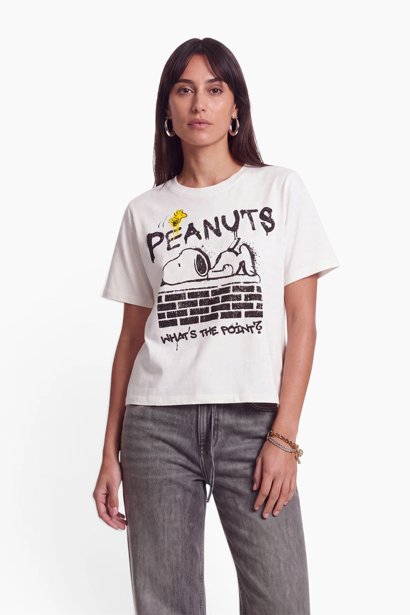 Eine Frau, die ein cremefarbenes „Peanuts“-T-Shirt trägt, auf dem Snoopy abgebildet ist, der auf einer Backsteinmauer liegt; darüber steht „PEANUTS“ und darunter „WHATS THE POINT?“.
