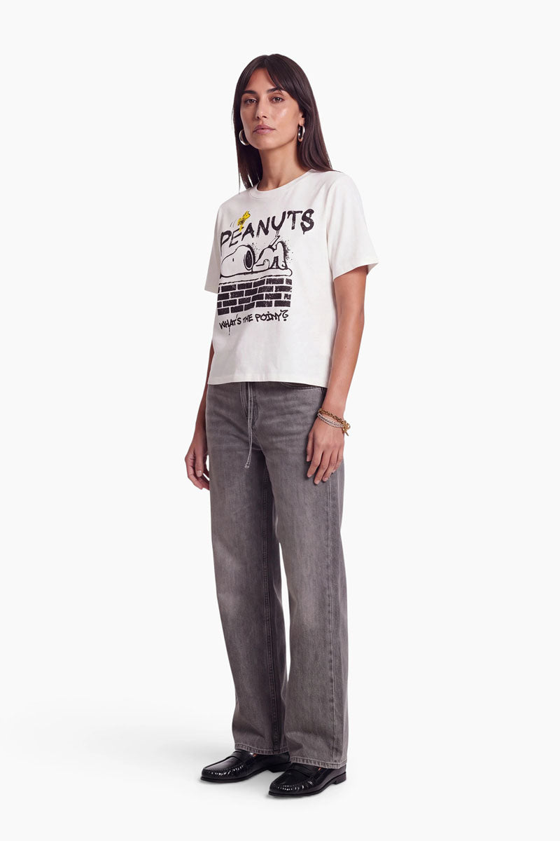 Eine Frau präsentiert ein weißes „Peanuts“-T-Shirt aus 100 % Baumwolle, auf dem Snoopy auf einer Backsteinmauer liegend abgebildet ist, zusammen mit dem Schriftzug „What’s the point?“. Sie trägt eine anthrazitfarbene Jeans mit weitem Bein und schwarze Loafer.