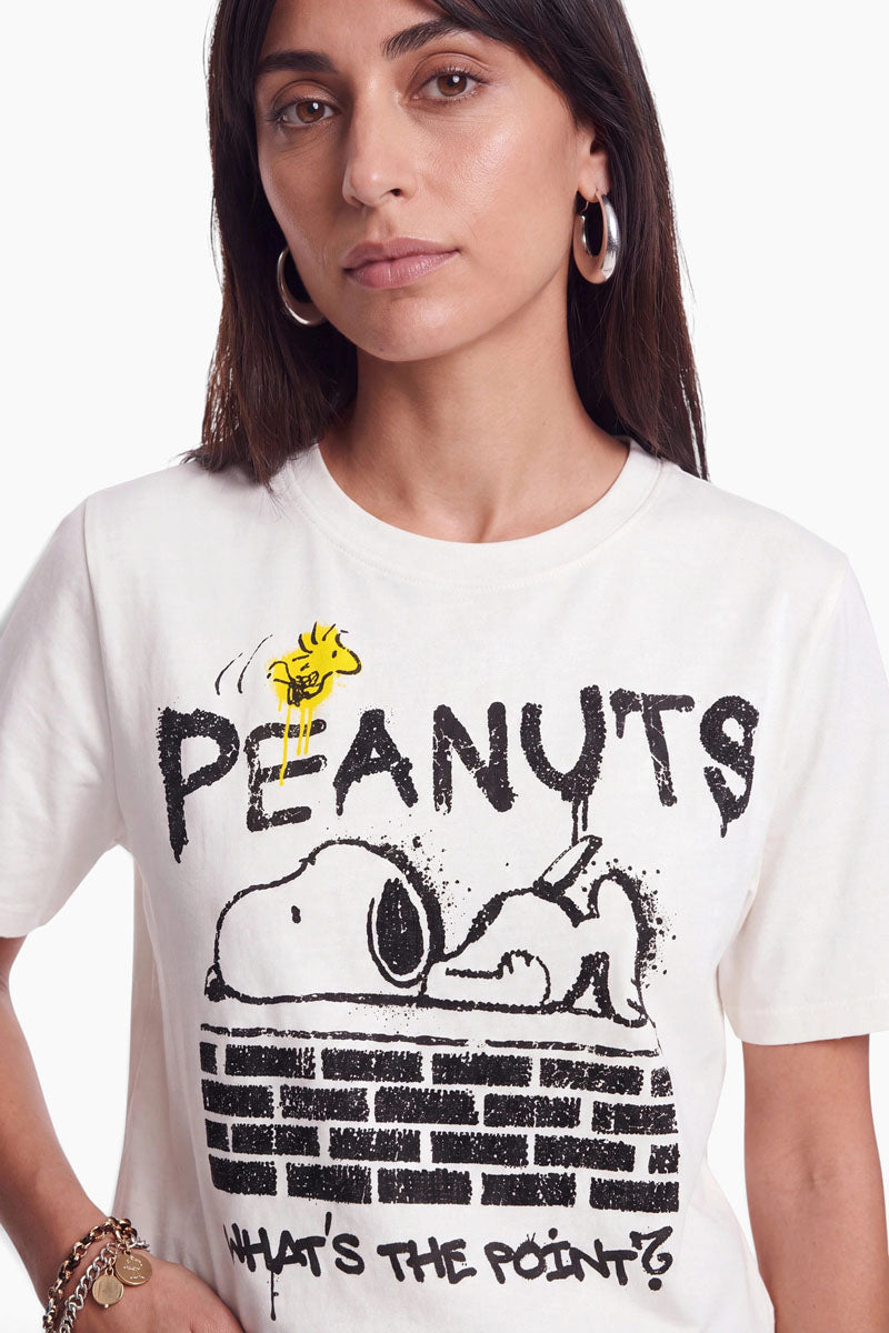 Eine Frau trägt ein cremefarbenes T-Shirt aus 100 % Baumwolle mit einem „Peanuts“-Motiv, auf dem Snoopy auf einer Backsteinmauer liegt; um die Illustration herum sind die Schriftzüge „PEANUTS“ und „WHATS THE POINT?“ aufgedruckt.