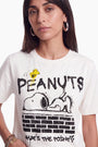 Eine Frau trägt ein cremefarbenes T-Shirt aus 100 % Baumwolle mit einem „Peanuts“-Motiv, auf dem Snoopy auf einer Backsteinmauer liegt; um die Illustration herum sind die Schriftzüge „PEANUTS“ und „WHATS THE POINT?“ aufgedruckt.