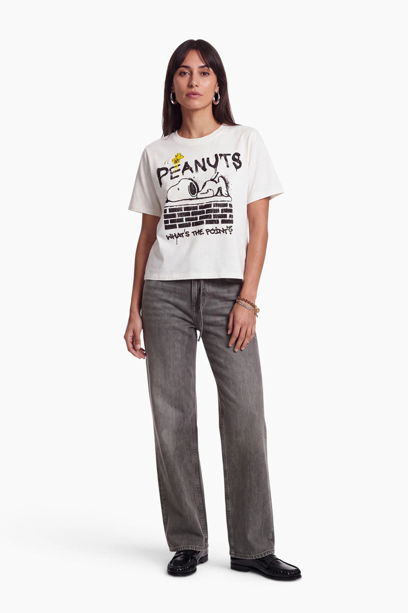 Eine Frau trägt ein weißes „Peanuts“-T-Shirt aus 100 % Baumwolle, auf dem Snoopy zu sehen ist, der auf einer Backsteinmauer liegt, sowie den Schriftzug „What’s the point?“. Außerdem trägt sie eine graue Jeans und schwarze Schuhe.
