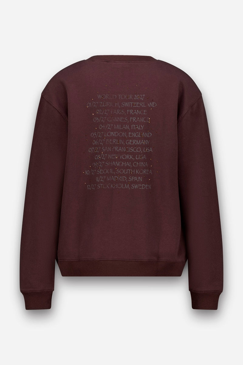 Rückansicht eines dunkelrotbraunen Baumwoll-Sweatshirts mit einer Stickerei, auf der eine „World Tour 2027“ mit verschiedenen Städten weltweit und den jeweiligen Terminen aufgeführt ist, beginnend mit Zürich im Januar und endend mit Stockholm im Dezember.
