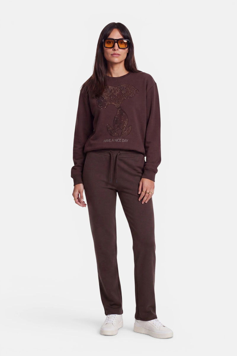 Eine Frau mit glattem braunem Haar trägt eine braune Sonnenbrille, ein dunkelbraunes Sweatshirt mit einem Snoopy-Motiv und dem Schriftzug „Have a Nice Day“, dazu passende braune Jogginghosen aus Baumwolle mit elastischem Bund sowie weiße Sneaker; sie steht vor einem weißen Hintergrund.