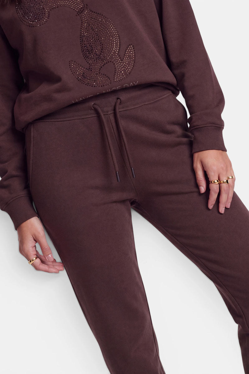 Eine Person, die einen braunen, zusammenpassenden Trainingsanzug aus weicher Baumwolle trägt, bestehend aus einer Trainingshose mit Kordelzug und elastischem Bund sowie einem Sweatshirt mit Strass-Motiv. Die Hand der Person ruht auf der Hüfte, an der mehrere goldene Ringe zu sehen sind. Der Hintergrund ist schlicht weiß. Zu sehen sind nur der Oberkörper und die Beine.