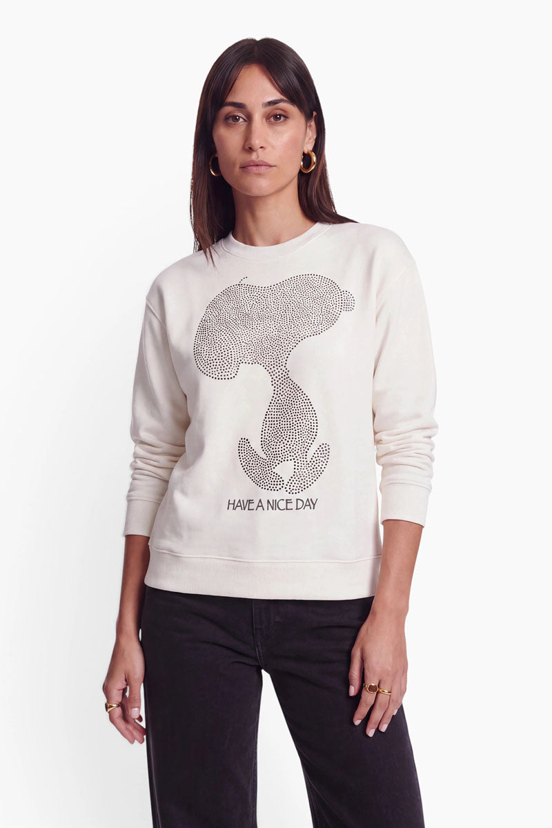Eine Frau trägt ein cremefarbenes Baumwoll-Sweatshirt mit einem großen Snoopy-Aufdruck aus schwarzen Nieten und dem Schriftzug „HAVE A NICE DAY.“ darunter.