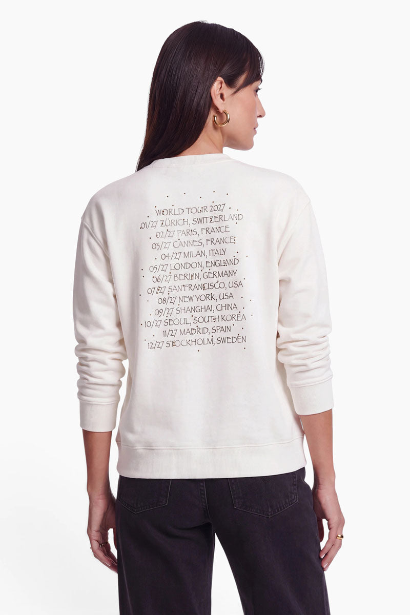 Eine Person, die ein cremefarbenes Baumwoll-Sweatshirt trägt, auf dessen Rücken ein Snoopy-Aufdruck sowie die Aufschrift „World Tour 2027“ neben einer Liste internationaler Städte aufgedruckt sind.
