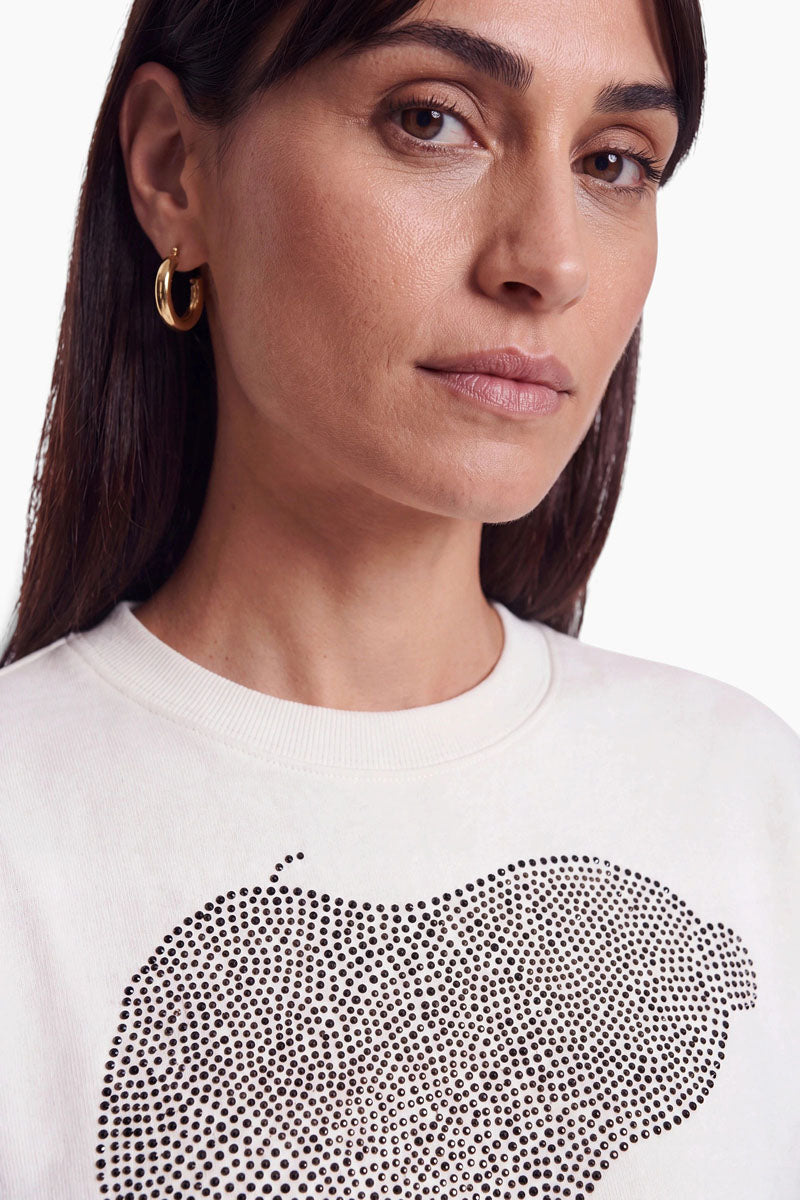 Eine Frau, die ein weißes Sweatshirt aus Baumwolle mit Rundhalsausschnitt trägt, auf dem ein großes grafisches Motiv aus kleinen schwarzen Nieten in Form einer Zitrone zu sehen ist.