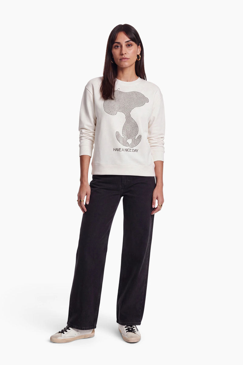 Eine Frau trägt ein cremefarbenes Baumwoll-Sweatshirt mit einem Snoopy-Motiv aus Punkten und dem Schriftzug „HAVE A NICE DAY“. Dazu trägt sie schwarze Jeans und lässige Sneaker.