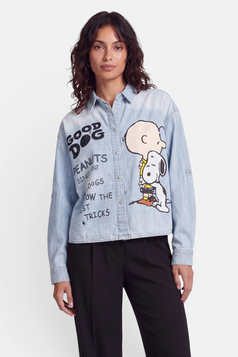 Eine Frau mit welligem, dunklem Haar trägt ein hellblaues Jeanshemd mit großen Abbildungen von Snoopy und Charlie Brown. Das Hemd ist mit verspielten Schriftzügen wie „GOOD DOG“, „PEANUTS“ und „SHOW THE DOGS THE BEST TRICKS“ verziert. Zu dem lustigen Oberteil trägt sie eine schwarze Hose.