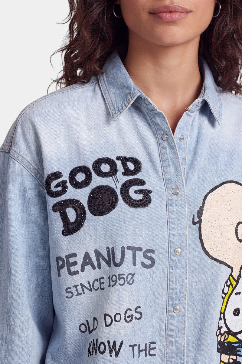 Eine Frau mit welligem braunem Haar trägt ein helles Jeanshemd mit der Aufschrift „GOOD DOG“ aus schwarzen Pailletten, dem Aufdruck „PEANUTS SINCE 1950“ und einer Figur, die Charlie Brown ähnelt, auf der rechten Seite. Das Hemd hat silberne Knöpfe mit Strassverzierung und einen spitzen Kragen, was eine verspielte Anspielung auf den ikonischen Snoopy- und Charlie-Brown-Stil darstellt.