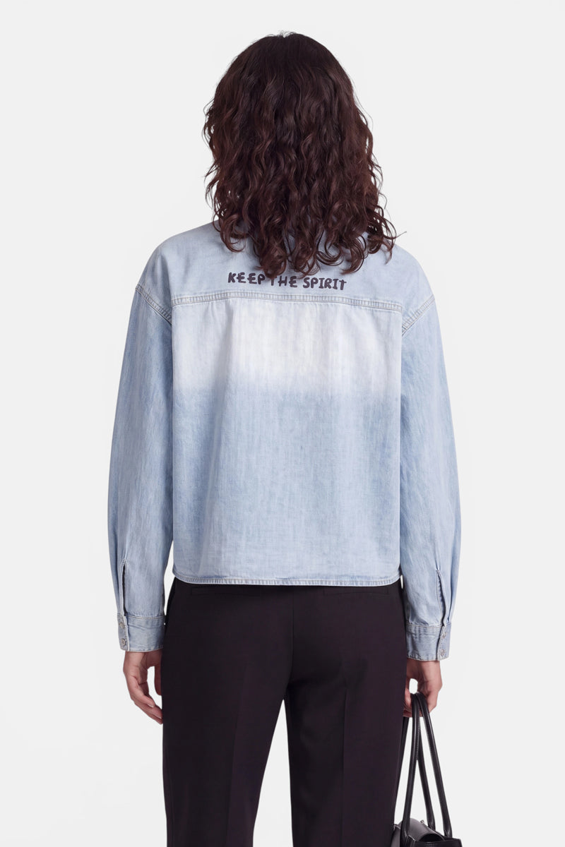 Eine Person mit schulterlangen Locken, von hinten gesehen, trägt ein helles Jeanshemd, auf dessen oberem Rücken in Strassakzenten „KEEP THE SPIRIT“ steht, sowie eine schwarze Hose. In der linken Hand hält sie eine schwarze Handtasche. Der Hintergrund ist schlicht weiß.