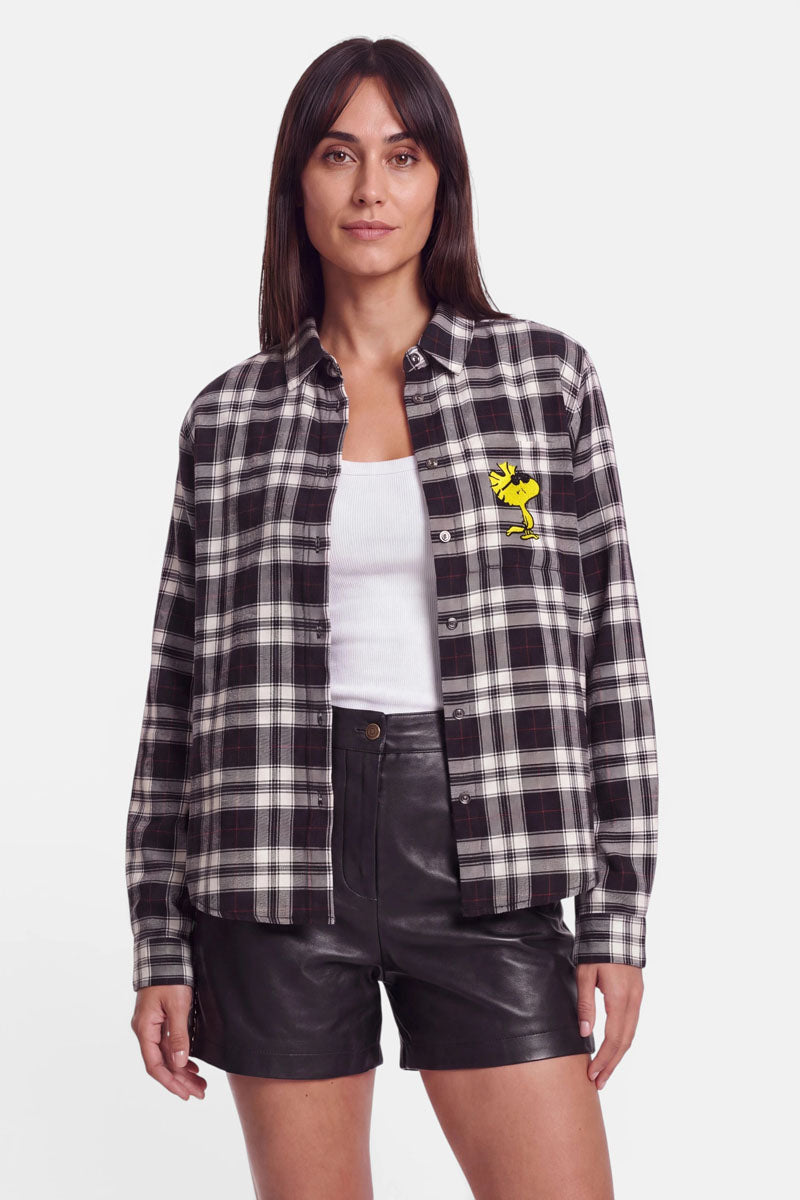 Eine Frau mit glattem, dunklem Haar trägt eine schwarz-weiße Karoprint-Hemdbluse mit einem gelben Snoopy-Aufnäher auf der Brust, dazu ein weißes Trägertop und schwarze Ledershorts. Sie steht mit dem Gesicht nach vorne vor einem schlichten weißen Hintergrund.