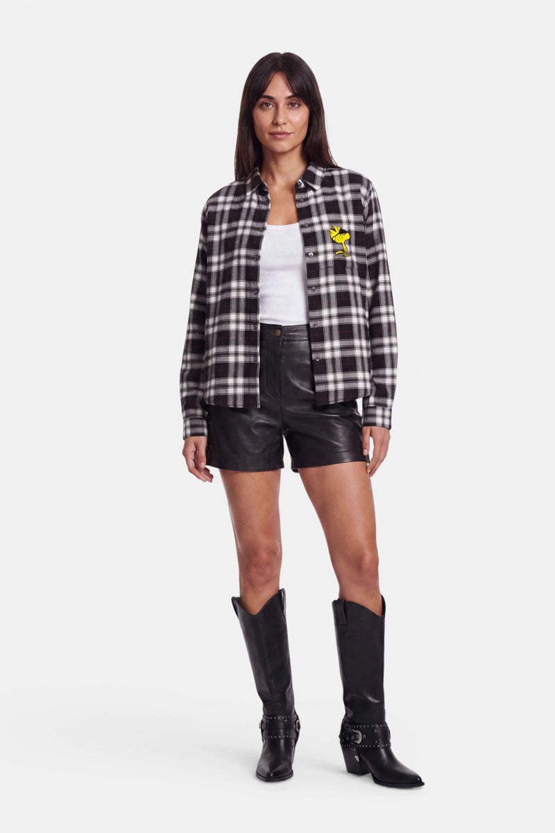 Eine Frau steht mit dem Gesicht nach vorne und trägt eine schwarz-weiße Karoprint-Hemdbluse mit einem gelben Vogel-Aufnäher, ein weißes Oberteil, schwarze Ledershorts und schwarze kniehohe Cowboystiefel. Sie hat glattes dunkles Haar, einen neutralen Gesichtsausdruck und posiert vor einem schlichten weißen Hintergrund.